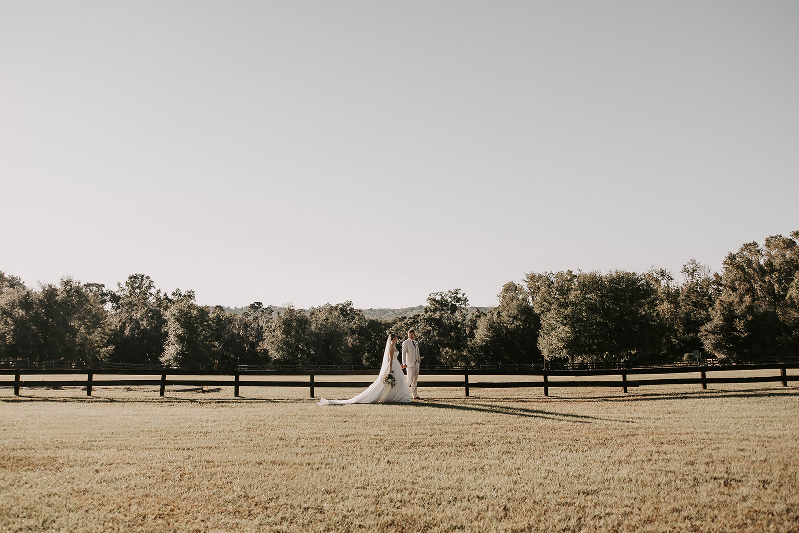bride groom portrait la hacienda snow hill brooksville florida - A Fall Wedding at La Hacienda on Snow Hill in Brooksville, FL Bride and groom standing together in an open field at La Hacienda on Snow Hill wedding venue in Brooksville Florida, with a long cathedral veil train and wood fence backdrop surrounded by trees