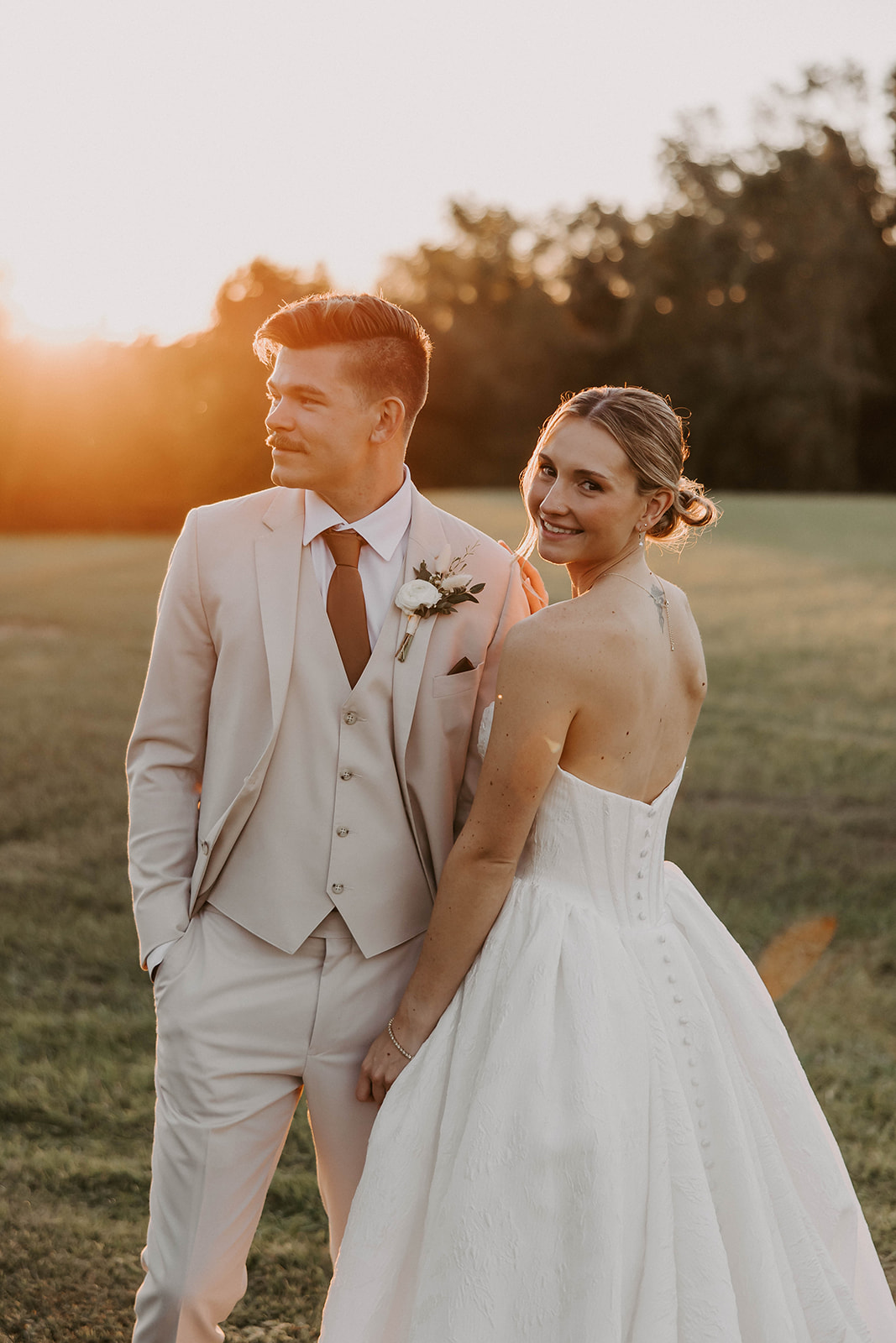 golden hour bride groom portrait la hacienda brooksville - A Fall Wedding at La Hacienda on Snow Hill in Brooksville, FL Bride in a backless button-detail gown looks over her shoulder smiling beside her groom in a tan three-piece suit during golden hour at La Hacienda on Snow Hill wedding venue in Brooksville Florida near Tampa